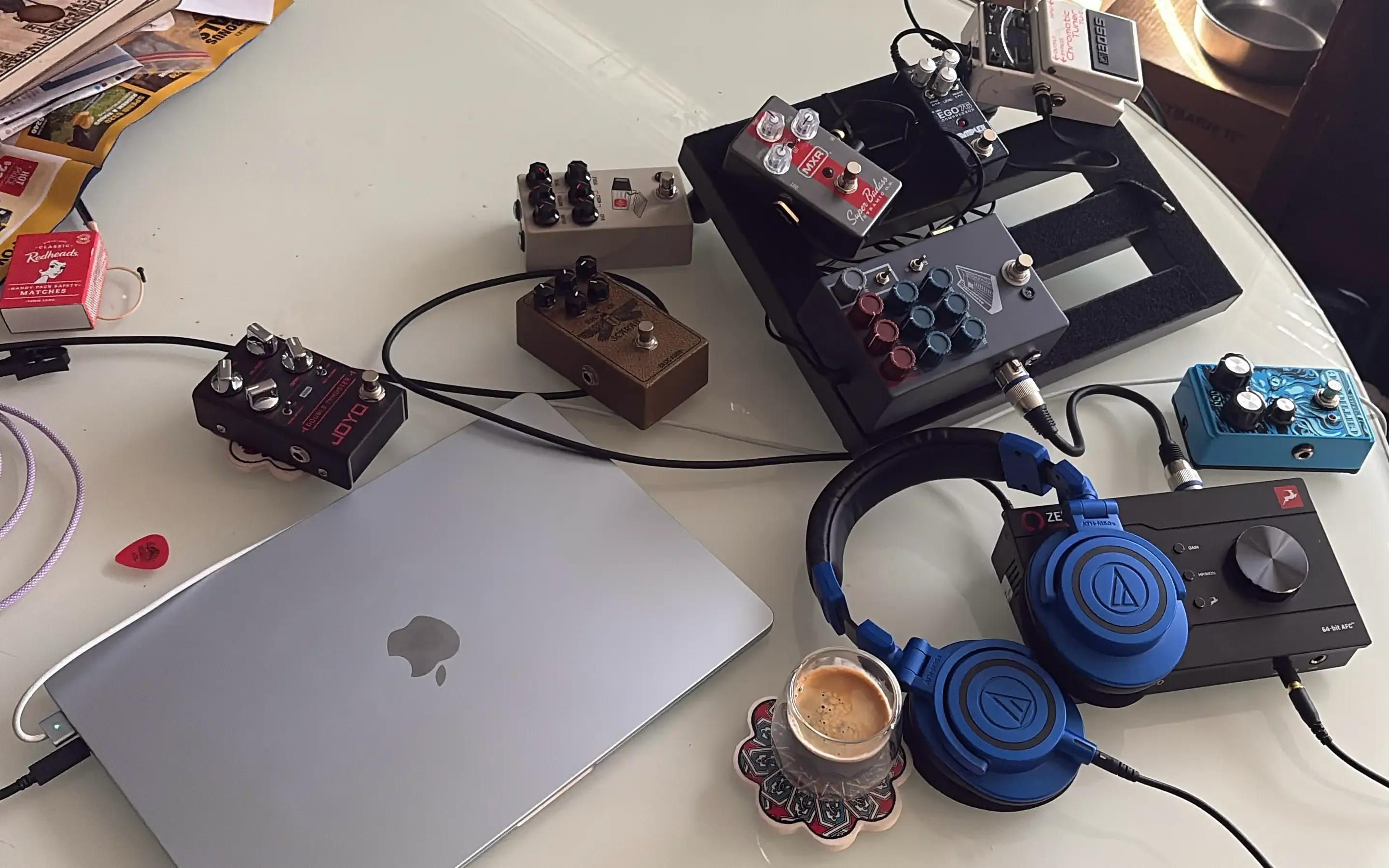 Pedalboard, MacBook, headphones and audio interface set up on a glass dining table, with a coffee on the side